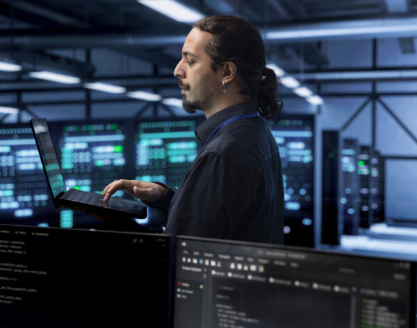 Engineer in data center using laptop to find firewall misconfigurations affecting rackmounts performance. Worker in server farm checking systems bottlenecks leading to sluggish data transfer rates