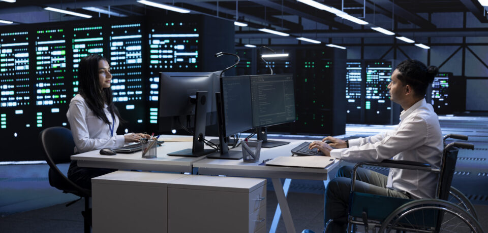 Person with paraplegia and colleague working in inclusive server hub workspace, typing on PC. Man in wheelchair and coworker doing tasks in data center housing advanced data storage infrastructure